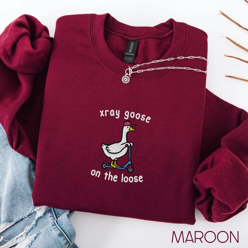 K&ouml;nnte beinhalten: Burgunderfarbenes Sweatshirt mit dem gestickten Schriftzug "xray goose on the loose" und einer Gans auf einem Roller. Eine silberne Halskette mit einem Sonnenanh&auml;nger liegt auf dem Sweatshirt. Das Wort "MAROON" steht unten im Bild.
