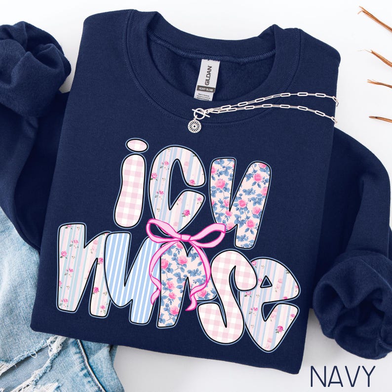 May include: Navy blue sweatshirt with the text "ICU Nurse" in a whimsical font. The letters are decorated with pink floral, gingham, and striped patterns. A pink bow is centered below the text. A silver necklace is draped across the sweatshirt.