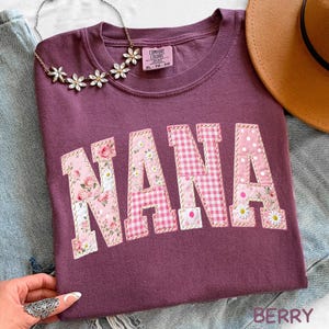 Peut inclure: T-shirt Comfort Colors de couleur baie avec le mot "NANA" appliqué en tissus à motifs floraux, vichy et à pois. Un collier floral et un chapeau marron sont visibles. Le t-shirt est de taille XL-TG.