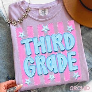 May include: A light purple t-shirt with the words "THIRD GRADE" in light blue, outlined in a darker blue. Pink vertical stripes and star accents decorate the shirt. The shirt is part of a back-to-school collection.