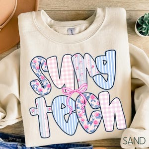 May include: A sand-colored sweatshirt with the words "SUNNY TEACH" in a bubble font. The letters are decorated with pink floral patterns, blue stripes, and pink gingham. A pink bow is between the words. The word "SAND" is in the bottom right corner.