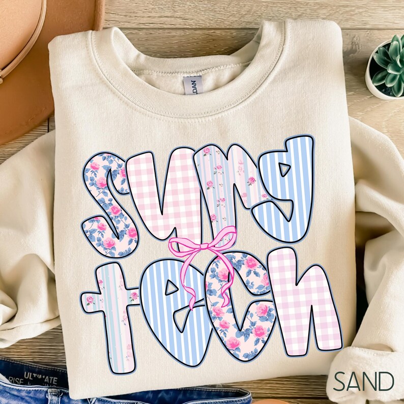 May include: A sand-colored sweatshirt with the words "SUNNY TEACH" in a bubble font. The letters are decorated with pink floral patterns, blue stripes, and pink gingham. A pink bow is between the words. The word "SAND" is in the bottom right corner.