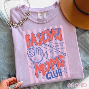 Op de afbeelding: Een lila Comfort Colors T-shirt met een honkbalthema. Het ontwerp toont de woorden "BASEBALL MOMS CLUB" in rood en blauw, samen met een honkbal, knuppel en handschoen. Het shirt heeft een relaxte pasvorm.