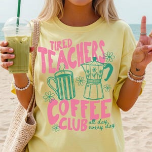 May include: A light yellow t-shirt with the words "Tired Teachers Coffee Club" in pink and green. The design includes a coffee cup, a coffee pot, and floral accents. The shirt is worn by a person holding a green drink.
