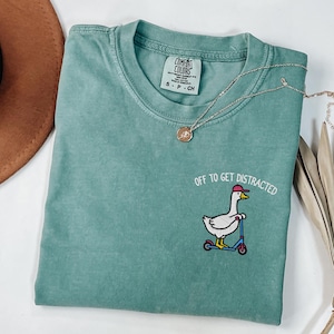 May include: A teal t-shirt featuring the embroidered text "OFF TO GET DISTRACTED" and a cartoon goose on a scooter. The shirt has a crew neck and short sleeves. A silver necklace with a pendant is draped across the shirt.