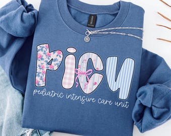 May include: Indigo blue sweatshirt with the word "PICU" in a whimsical font, featuring floral and striped patterns. The text "pediatric intensive care unit" is below the word. A silver necklace with a small pendant is draped across the sweatshirt.