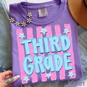 May include: Violet-colored t-shirt with the words "THIRD GRADE" in light blue, outlined in a darker blue, and set against pink vertical stripes. White star accents are scattered around the text. The shirt is folded.