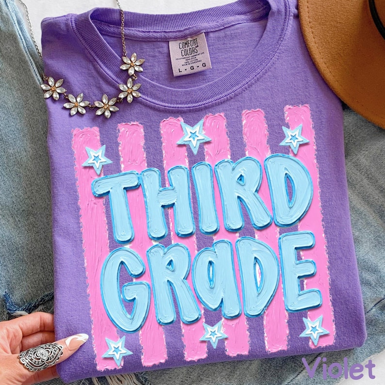 May include: Violet-colored t-shirt with the words "THIRD GRADE" in light blue, outlined in a darker blue, and set against pink vertical stripes. White star accents are scattered around the text. The shirt is folded.