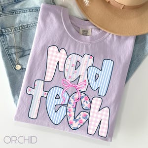 May include: A lilac t-shirt with the words "Mad Tech" in a playful font. The letters feature pink gingham, blue stripes, and floral patterns, with a pink bow accent. The word "ORCHID" is printed at the bottom.