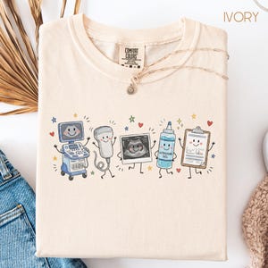 Peut inclure: Un t-shirt ivoire avec une impression graphique représentant cinq objets médicaux anthropomorphes : une machine à échographie, une sonde, une image d'échographie, un flacon de gel et un presse-papiers, entourés de petites étoiles et cœurs.