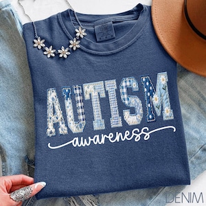 Peut inclure: Un t-shirt bleu marine avec les mots "AUTISM awareness" dans un motif patchwork. Les lettres sont dans divers motifs bleus, dont floral et vichy. Un collier chaîne de marguerites argentées et un chapeau marron sont également visibles.