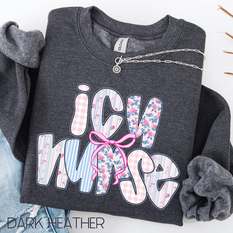May include: Dark heather gray sweatshirt with the text "ICU Nurse" in a bubble font. The letters are decorated with pink gingham, blue stripes, and floral patterns. A pink bow is centered below the letters. A silver necklace is draped across the sweatshirt.