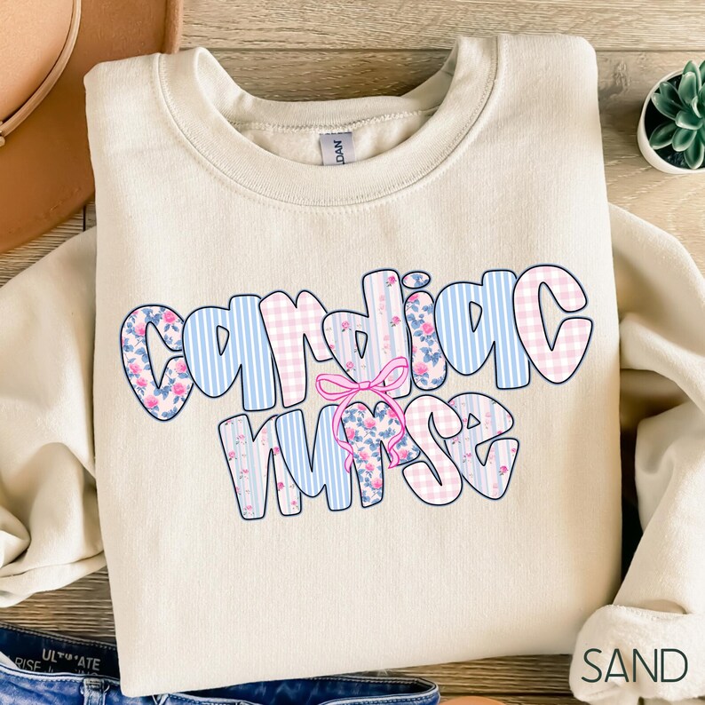 K&ouml;nnte beinhalten: Ein sandfarbenes Sweatshirt mit dem Schriftzug "Cardiac Nurse" in einer Blasen-Schriftart. Die Buchstaben sind mit blauen Streifen, rosa Blumenmustern und rosa Vichy-Karo verziert. Zwischen den W&ouml;rtern befindet sich eine rosa Schleife.