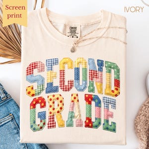 May include: Ivory-colored t-shirt with the words "SECOND GRADE" in colorful patchwork letters. The shirt has a crew neck and long sleeves. A gold necklace with a pendant is draped on the shirt. The words "Screen print" are in a yellow box.