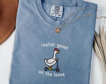 May include: A blue jean colored t-shirt with the words "realtor goose on the loose" embroidered on the front. The graphic shows a goose riding a scooter and wearing a hat. The shirt has a Comfort Colors tag.