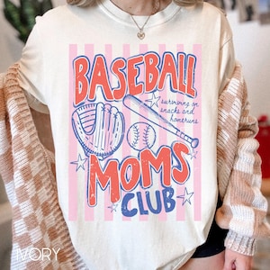Op de afbeelding: Cr&egrave;mekleurig T-shirt met een honkbalthema. Het ontwerp toont de woorden "BASEBALL MOMS CLUB" in rood en blauw, een honkbalhandschoen, een knuppel en een bal. Roze en witte strepen vormen de achtergrond.