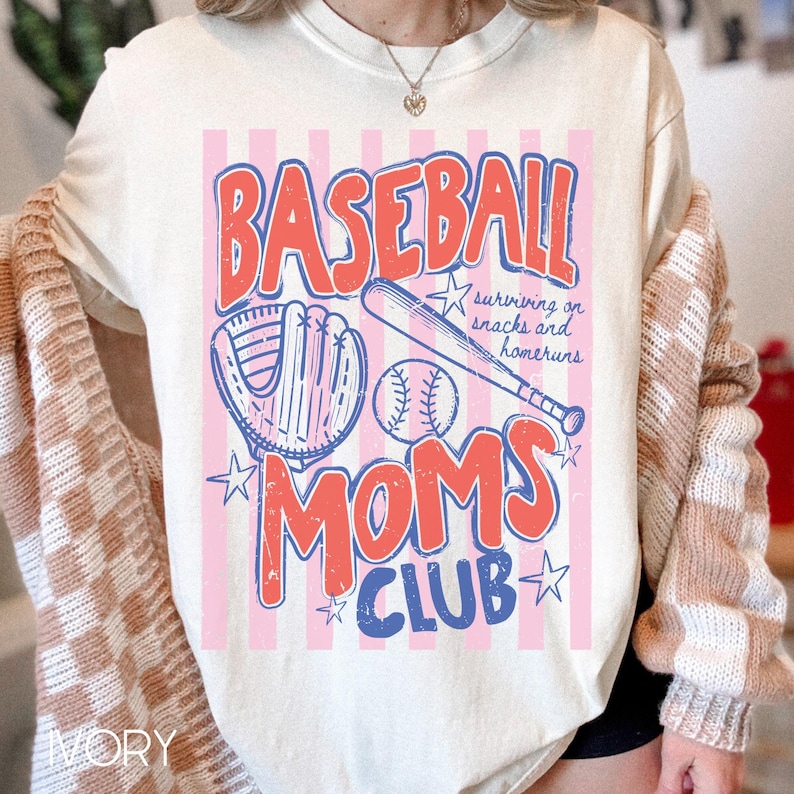 Op de afbeelding: Cr&egrave;mekleurig T-shirt met een honkbalthema. Het ontwerp toont de woorden "BASEBALL MOMS CLUB" in rood en blauw, een honkbalhandschoen, een knuppel en een bal. Roze en witte strepen vormen de achtergrond.