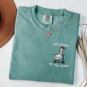 May include: A teal Comfort Colors t-shirt with the phrase "pta goose on the loose" and an embroidered goose wearing a hat and riding a scooter. A silver necklace with a pendant is on the shirt.