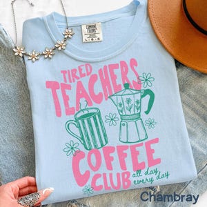 May include: Light blue t-shirt with the text "Tired Teachers Coffee Club" in pink and green. The design includes a coffee cup, a coffee pot, and flowers. The shirt is a Comfort Colors brand.