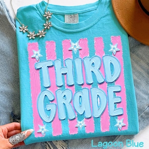 May include: A Lagoon Blue t-shirt with the words "THIRD GRADE" in light blue, outlined in a darker blue. The design includes pink vertical stripes and white star accents. A floral necklace and a brown hat are also visible.