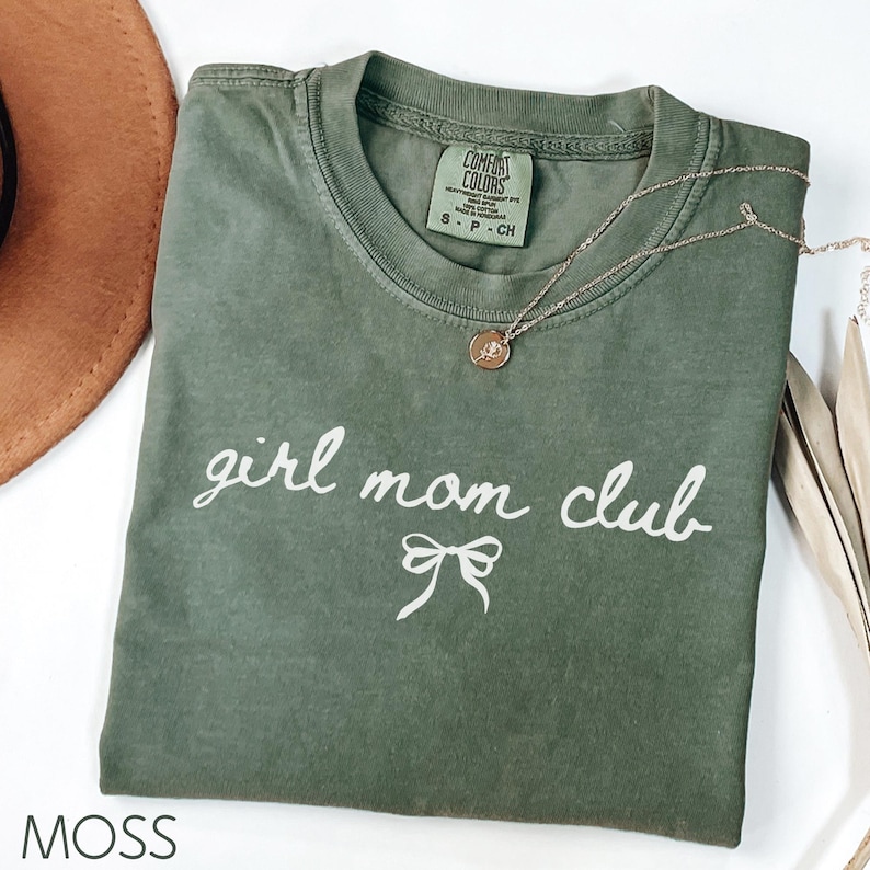 Peut inclure: Un t-shirt vert avec le texte "girl mom club" en &eacute;criture blanche avec un n&oelig;ud blanc en dessous. Le t-shirt est pli&eacute; et pos&eacute; &agrave; plat sur une surface blanche. Le mot "MOSS" est imprim&eacute; en blanc en bas de l'image.