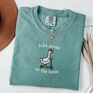 May include: A teal Comfort Colors t-shirt with the embroidered phrase "bcba goose on the loose" and a cartoon goose on a scooter. A silver necklace with a pendant is draped across the shirt.