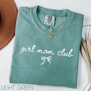 Peut inclure: T-shirt vert clair avec le texte "girl mom club" en lettres blanches et un n&oelig;ud blanc. Le t-shirt a un aspect vieilli et est fait d'un tissu doux et confortable.