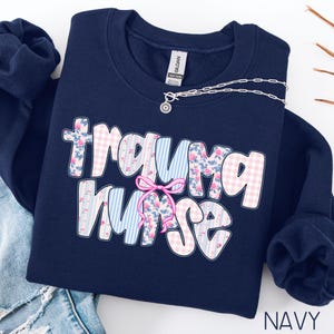 Puede incluir: Sudadera azul marino con la frase "Trauma Nurse" en un dise&ntilde;o floral y de cuadros. Un lazo rosa est&aacute; centrado en el dise&ntilde;o. Un collar plateado est&aacute; drapeado sobre la sudadera. La palabra "NAVY" est&aacute; impresa en la parte inferior derecha.