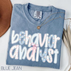 May include: A blue jean Comfort Colors t-shirt with the words "behavior analyst" in a decorative font. The letters are patterned with bows, stripes, and floral designs. A gold necklace with a pendant rests on the shirt.