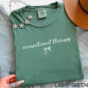 May include: Light green t-shirt with the words "occupational therapy" in white script and a bow graphic. The shirt is folded and has a Comfort Colors tag. A floral necklace and a brown hat are also visible.