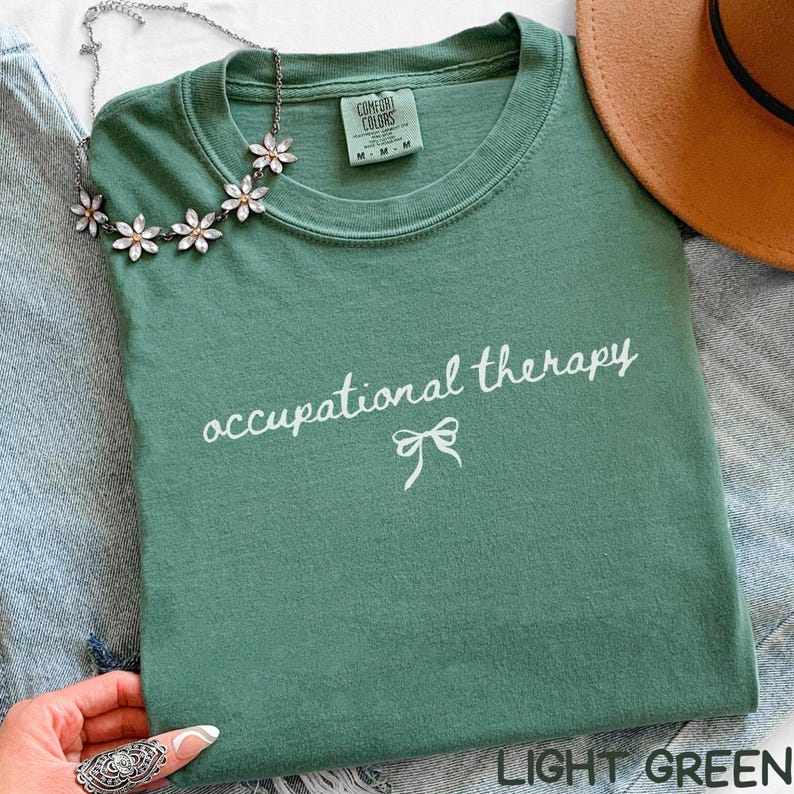 May include: Light green t-shirt with the words "occupational therapy" in white script and a bow graphic. The shirt is folded and has a Comfort Colors tag. A floral necklace and a brown hat are also visible.