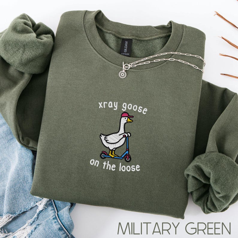 K&ouml;nnte beinhalten: Milit&auml;rgr&uuml;nes Sweatshirt mit dem gestickten Schriftzug "xray goose on the loose" und einer Cartoon-Gans auf einem Roller. Eine silberne Halskette mit Sonnenanh&auml;nger liegt auf dem Sweatshirt. Das Sweatshirt ist auf einer Jeans gefaltet.