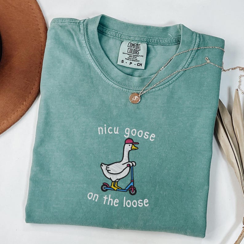 Peut inclure: Un t-shirt Comfort Colors vert d'eau avec l'inscription brod&eacute;e "nicu goose on the loose" et une oie sur un scooter. Le t-shirt a un col rond et des manches courtes. Un collier en or avec un pendentif est drap&eacute; sur le t-shirt.