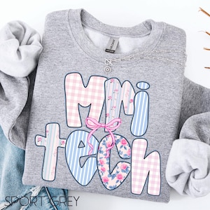May include: A gray sweatshirt with the words "Mini Teach" in a colorful design. The letters are decorated with pink gingham, blue stripes, and floral patterns. A pink bow is between the letters "i" and "t". A silver necklace is visible.