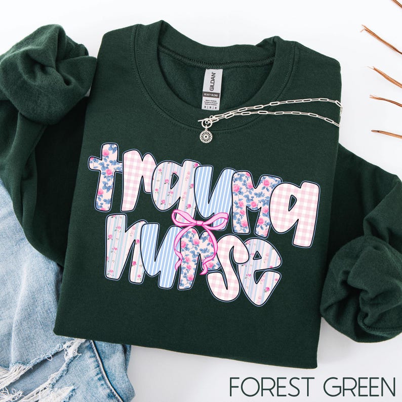 Puede incluir: Una sudadera verde oscuro con la frase "Trauma Nurse" en una fuente caprichosa. Las letras est&aacute;n decoradas con una mezcla de cuadros rosas, rayas azules y estampados florales. Tambi&eacute;n se ve un collar plateado con un colgante de sol.
