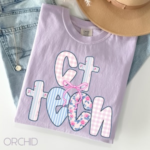 May include: A lilac-colored t-shirt with the words "CT TECH" in a whimsical font. The letters feature various patterns, including gingham, stripes, and floral designs, with a pink bow accent. The shirt is folded on top of blue jeans.