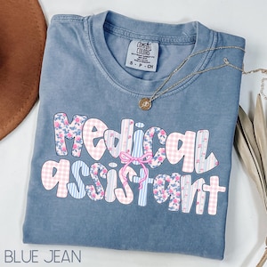 May include: A blue jean coloured t-shirt featuring the words "Medical Assistant" in a floral and gingham design. A gold necklace with a pendant is draped across the shirt. The shirt is folded and has a Comfort Colors tag.