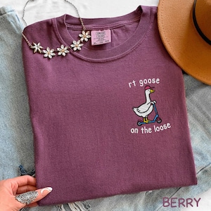 May include: Berry-colored t-shirt with the phrase "rt goose on the loose" and a cartoon goose riding a scooter. The shirt has a crew neck and short sleeves. A floral necklace and a brown hat are also visible.