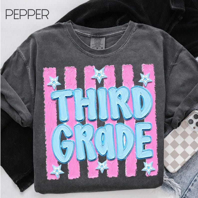 May include: Dark gray sweatshirt with the text "THIRD GRADE" in light blue, outlined in white, and set against a pink striped background. White star accents are scattered around the text. The word "PEPPER" is in the top left corner.