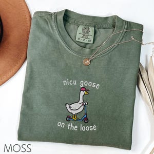Peut inclure: T-shirt vert mousse Comfort Colors avec la phrase brod&eacute;e "nicu goose on the loose" et un dessin d'oie sur un scooter. Un collier dor&eacute; et un chapeau marron sont &eacute;galement visibles.
