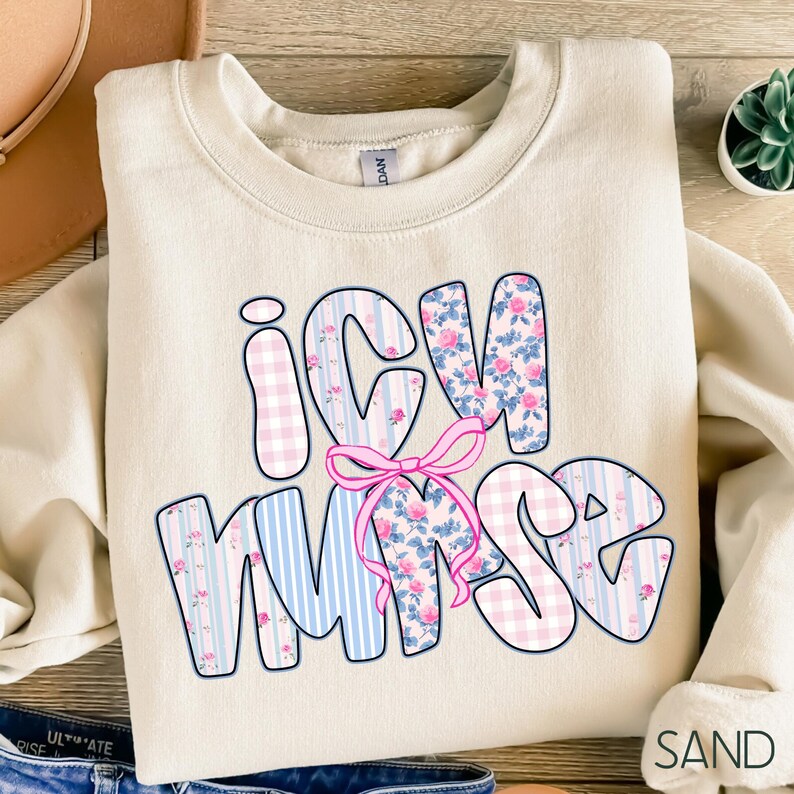 May include: A sand-colored sweatshirt with the text "ICU NURSE" in a bubble font. The letters are decorated with pink floral patterns, blue stripes, and pink gingham. A pink bow is centered between the letters. The sweatshirt is folded.