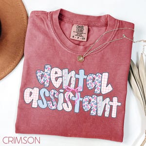 Pode incluir: Camiseta cor carmesim com a frase "Dental Assistant" em uma fonte divertida. As letras s&atilde;o decoradas com estampas florais, xadrez e listras. Um colar delicado com um pingente repousa sobre a camiseta.