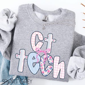 May include: A light gray sweatshirt with the words "Ct Tech" in a colorful design. The letters are decorated with pink gingham, blue stripes, and floral patterns. A pink bow accents the design. A silver necklace is visible.