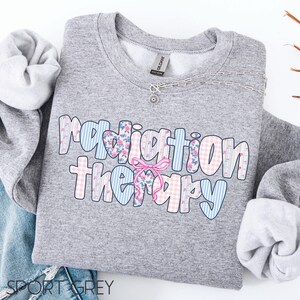 Może przedstawiać: Szara bluza z napisem "Radiation Therapy" w kolorowym, wzorzystym designie. Tekst zawiera niebieskie paski, r&oacute;żową kratę i kwiatowe wzory, z r&oacute;żową kokardą. Srebrny naszyjnik z małą zawieszką jest przewieszony przez bluzę.