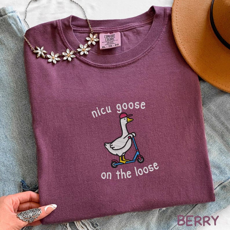 Peut inclure: T-shirt de couleur baie avec l'inscription "nicu goose on the loose" et un dessin de canard sur un scooter. Le t-shirt a un col rond et une &eacute;tiquette Comfort Colors. Un collier floral et un chapeau marron sont &eacute;galement visibles.