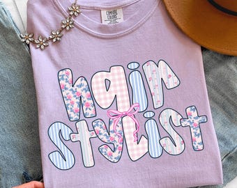 May include: A lilac Comfort Colors t-shirt with the words "hair stylist" in a bubble font. The letters are decorated with floral, gingham, and striped patterns. A pink bow is between the words. A necklace and a hat are in the background.