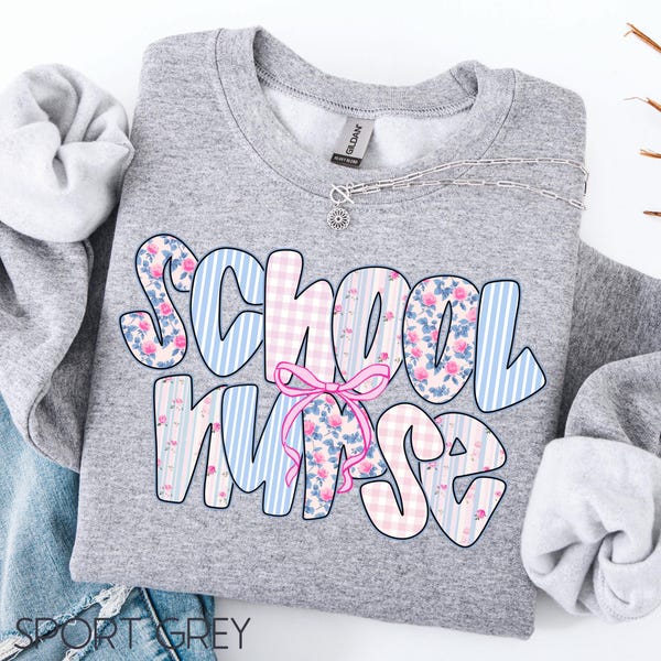 May include: A gray sweatshirt with the words "School Nurse" in a floral and striped design. The letters are in blue and pink, with a pink bow in the center. A silver necklace is visible at the top.