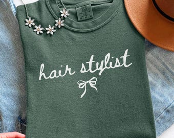 May include: A moss green sweatshirt with the words "hair stylist" in white script and a bow graphic. A silver necklace with flower-shaped pendants and a brown hat are also visible. The sweatshirt is folded on a denim surface.