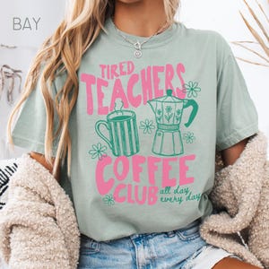 May include: A light green t-shirt with the text "Tired Teachers Coffee Club" in pink. The design includes a green coffee mug, a coffee pot, and flowers. The words "all day, every day" are also included in the design.