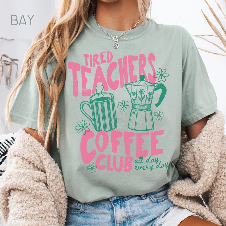 May include: A light green t-shirt with the text "Tired Teachers Coffee Club" in pink. The design includes a green coffee mug, a coffee pot, and flowers. The words "all day, every day" are also included in the design.